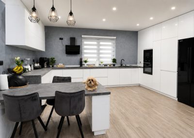 Bucătărie albă în L cu peninsulă pentru servit masa, blat imitație beton, pendule negre geometrice și frigider side-by-side