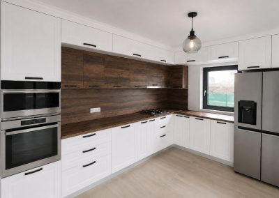 Bucătărie albă în L făcută la comandă, cu peretele placat în lemn de nuc, blat maro închis și coloană de electrocasnice inox