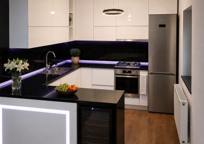 Bucătărie albă lucioasă în U cu bar iluminat, blat negru și bandă LED violet sub corpuri