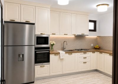 Bucatarie crem clasica in L cu corpuri pana in tavan, blat de lemn, frigider inox si coloana de cuptoare