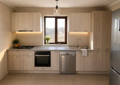 Bucatarie liniara cu fronturi imitatie stejar deschis, blat gri, fereastra centrala si masina de spalat vase inox
